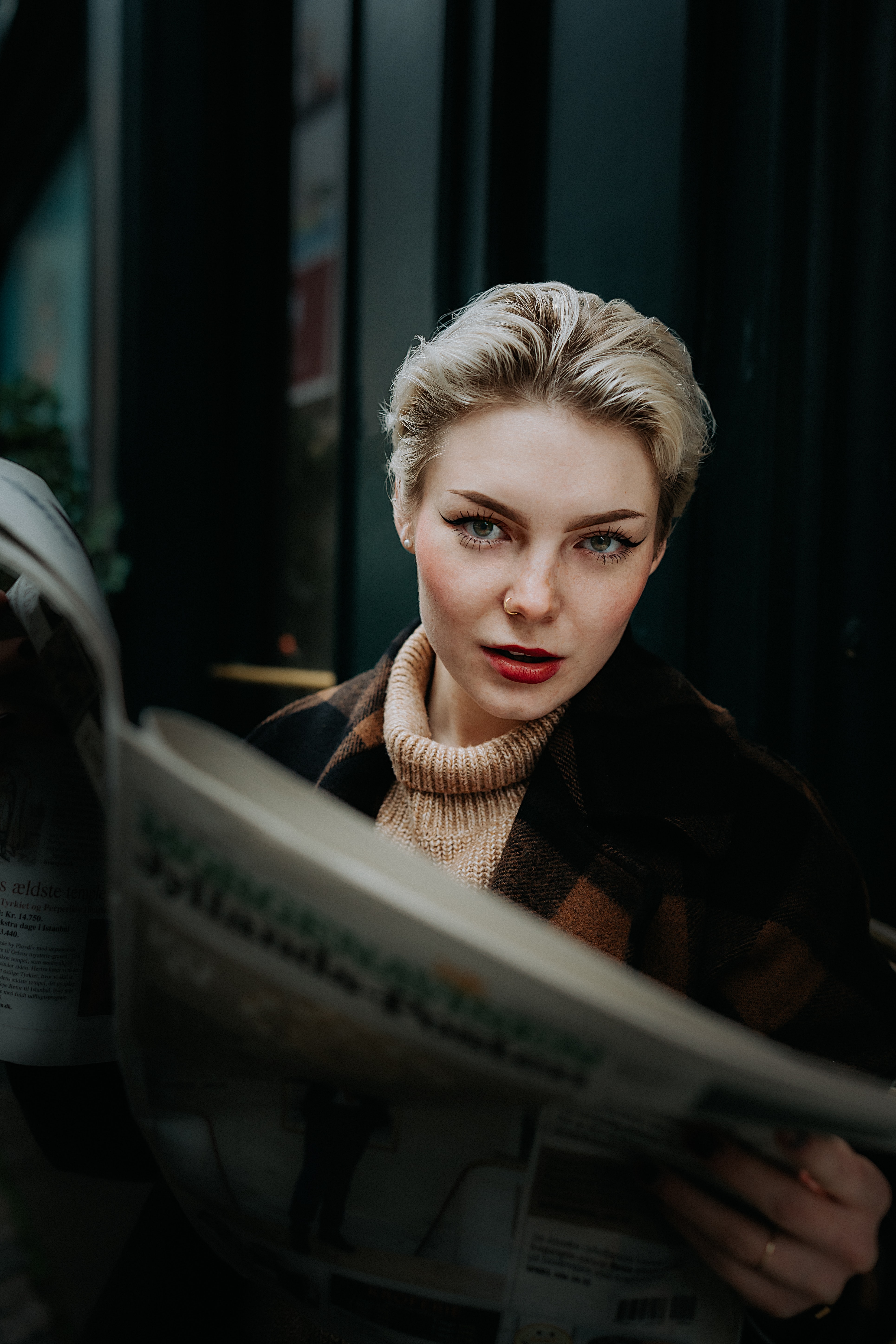 A beautiful, sophisticated woman with short blonde hair , darker roots, striking blue eyes, full eyebrows, and subtle red lipstick looking over the open full-size newspaper wearing a light brown turtle neck sweater. She appears to be sitting on darken subway bench seat however her face is well lite. She has a look as if she is waiting for an answer to a serious question she asked to the person in the perspective of the camera lens. 